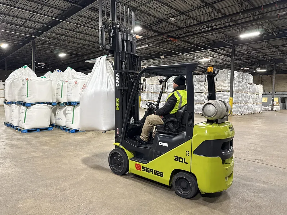 Yellow forklift transports large white bulk bags inside a spacious warehouse, demonstrating All City Leasing’s palletizing and material-handling capability.