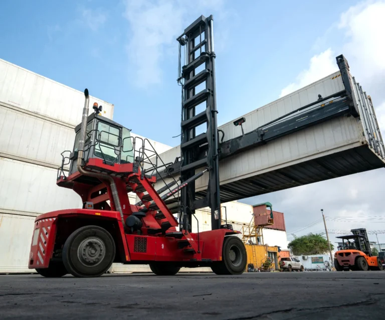Red container handler forklift lifting white shipping container at logistics yard with stacked units and cargo transport equipment in background