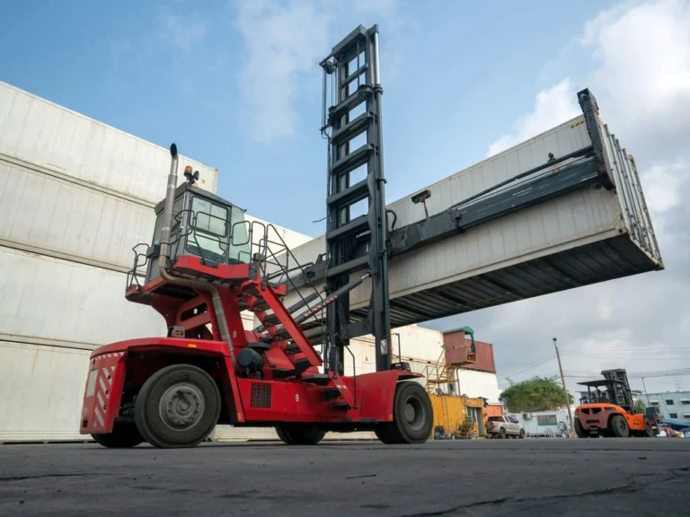 Large red forklift moving a white shipping container in an outdoor freight yard with other containers and equipment visible