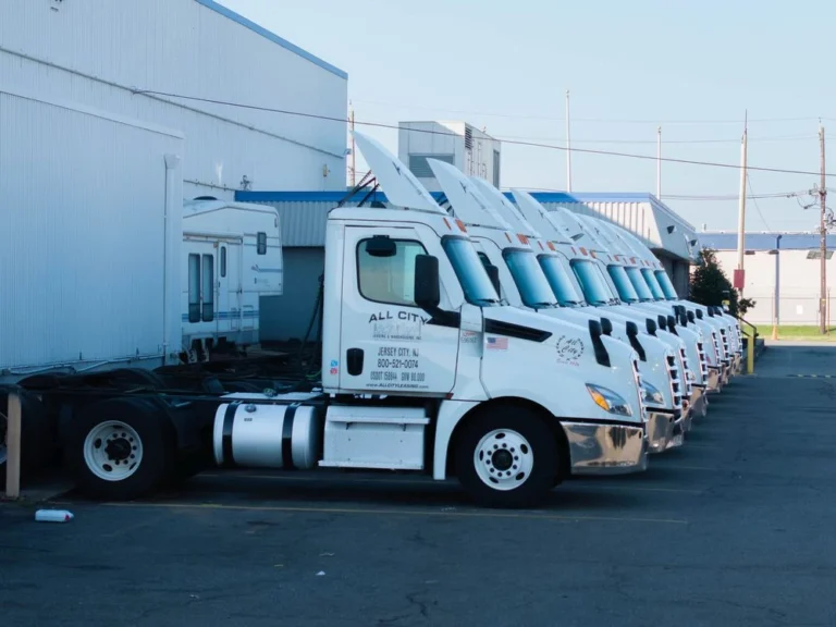 Line of white All City Leasing semi trucks parked side by side next to a warehouse in an industrial area