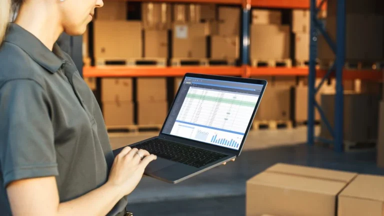Female warehouse employee using laptop with inventory spreadsheet open while standing among stacked cardboard boxes on storage shelves