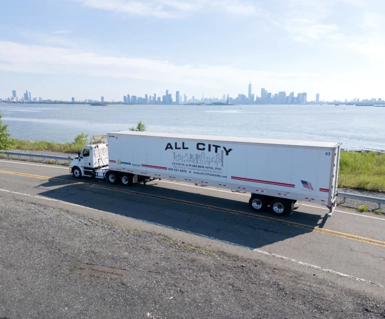 All City logistics semi truck driving along waterfront road with New York City skyline in background, providing freight and shipping services in the region